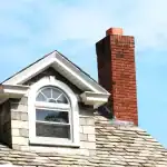 A close-up of a house roof with gray shingles, a red brick chimney, and a dormer window with white trim and an arched top, set against a blue sky—perfect for showcasing the need for air duct services New Jersey homeowners trust.