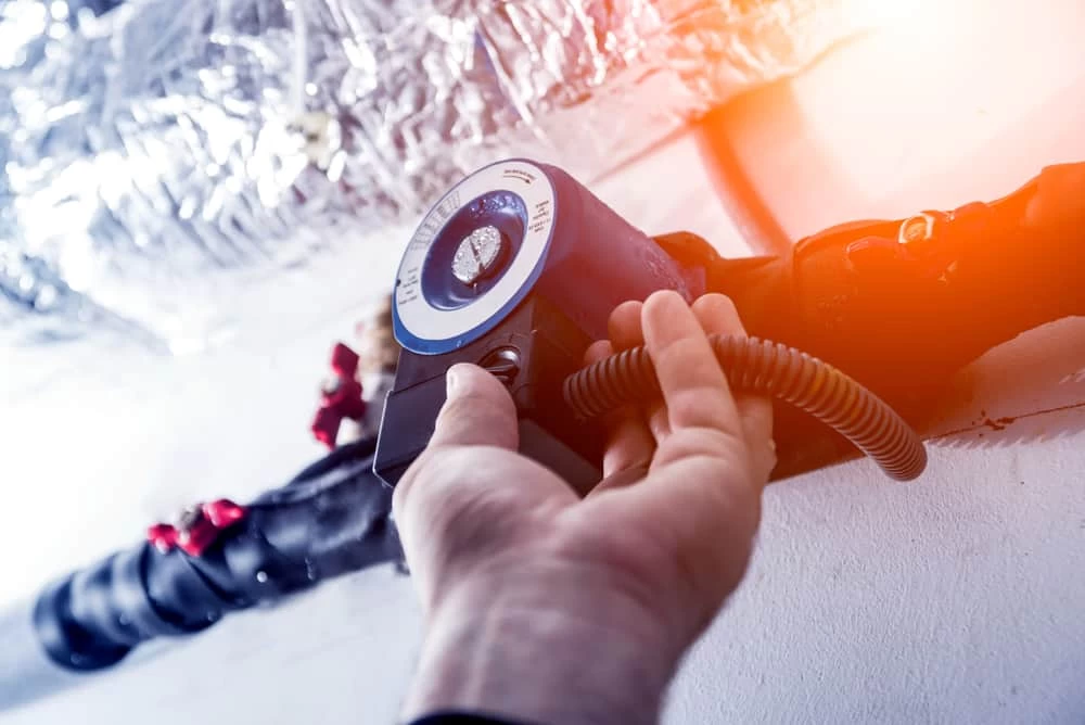 A hand adjusts a circular control device attached to insulated pipes on a ceiling—foil insulation gleams in the background and warm light shines from the right, highlighting routine air duct services New Jersey professionals might perform.