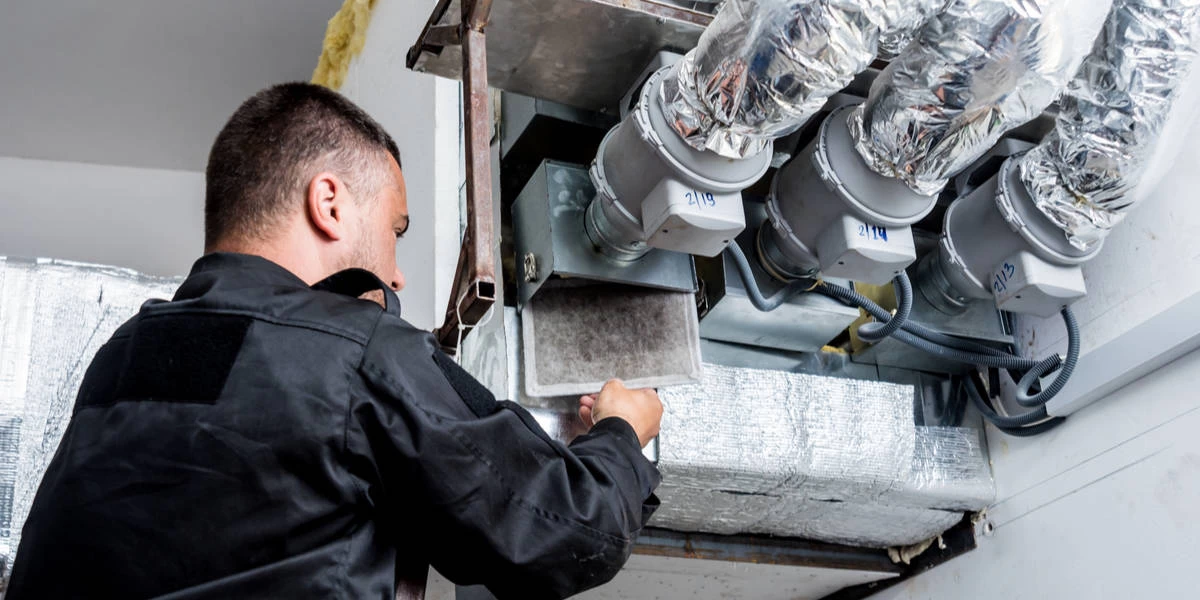 A worker in a black uniform inspects and maintains an HVAC ventilation system, adjusting or replacing a filter near large insulated air ducts as part of professional air duct services New Jersey.