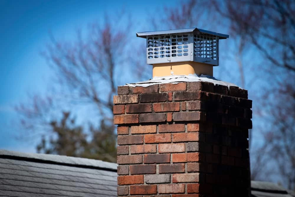 A brick chimney with a metal cap sits on a shingled roof, framed by bare tree branches and a clear blue sky—reminding homeowners of the importance of air duct cleaning New Jersey for fresh, healthy indoor air.