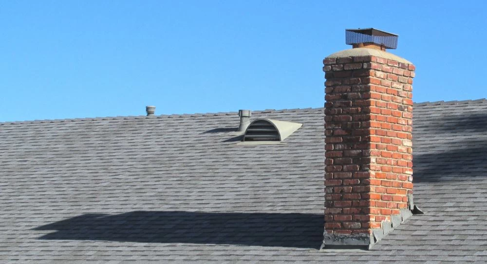 A red brick chimney rises from a gray shingled roof under a clear blue sky, with a metal vent and small exhaust pipe—ideal spots for dryer vent cleaning New Jersey professionals to inspect—also visible on the roof.