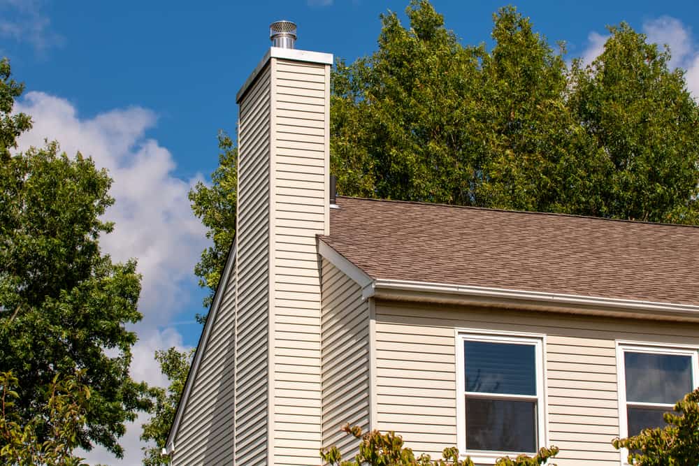 A beige house with tan siding and a tall chimney stands among green trees under a blue sky. Two windows are visible on the side—perfect for homeowners seeking air duct cleaning New Jersey to keep indoor air fresh and healthy.