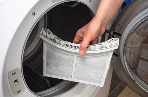 A person’s hand removes a lint filter covered in dust from the open door of a front-loading dryer, emphasizing the importance of regular dryer vent cleaning New Jersey to maintain safety and efficiency.