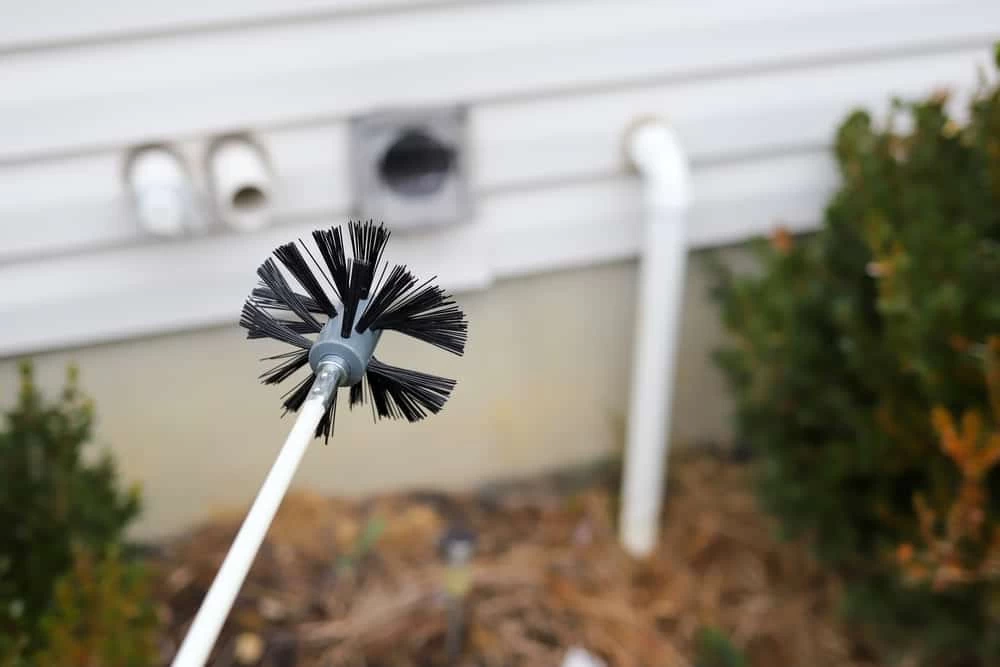 A close-up of a round dryer vent cleaning brush held outside, with a house wall, dryer vent, and some bushes visible in the background—perfect for showcasing expert dryer vent cleaning New Jersey services.
