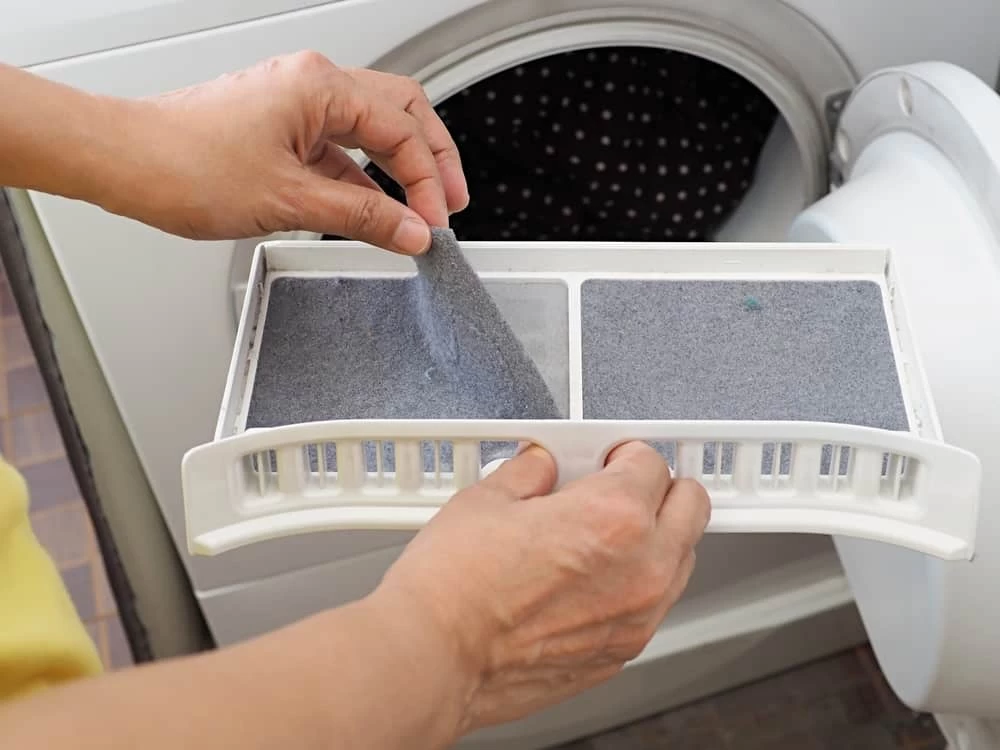 A person is removing lint from a filter tray of a front-loading dryer, with the dryer door open and clothes visible inside—essential for safe operation and recommended by dryer vent cleaning New Jersey professionals.