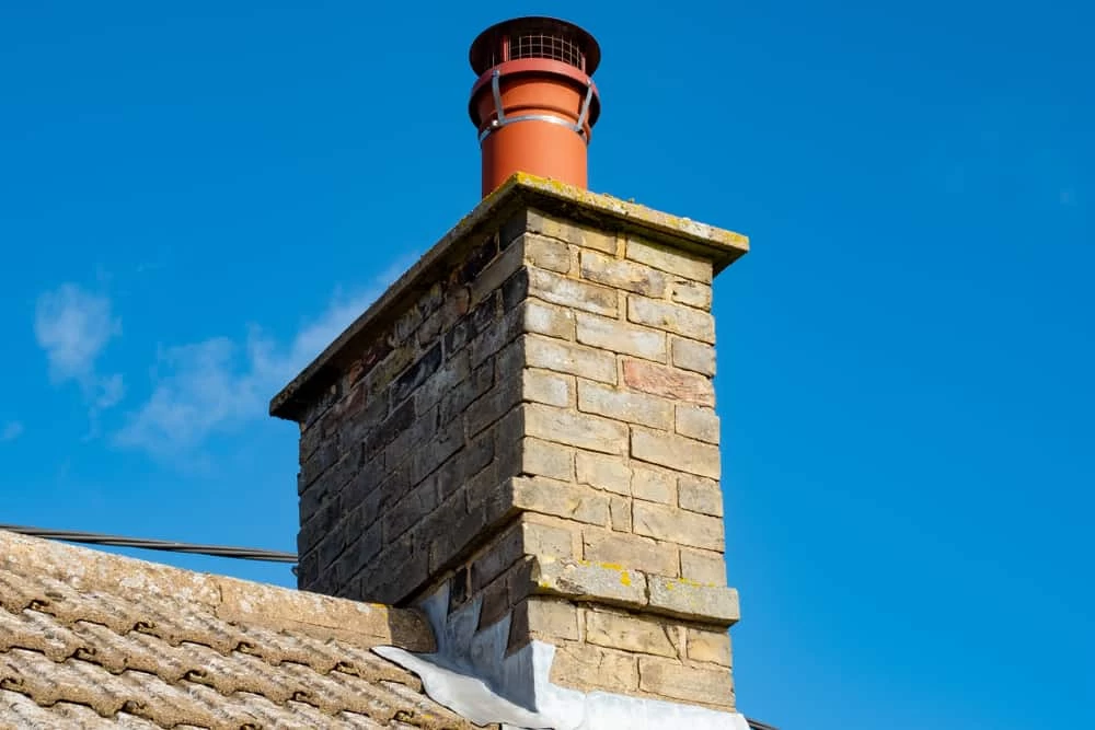 A close-up of a brick chimney with a red metal cap, set against a clear blue sky, on the roof of a house with gray shingles—ideal for showcasing expert dryer vent cleaning New Jersey homeowners can trust.
