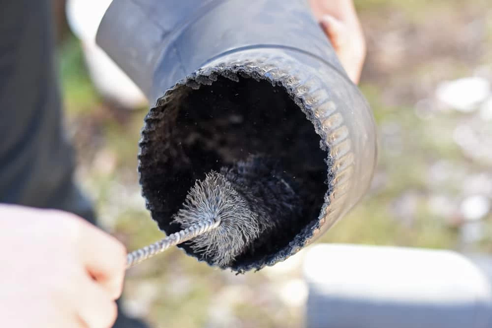 A person cleans the inside of a large black pipe with a round wire brush tool, focusing on removing soot or debris—similar to professional dryer vent cleaning New Jersey specialists provide.