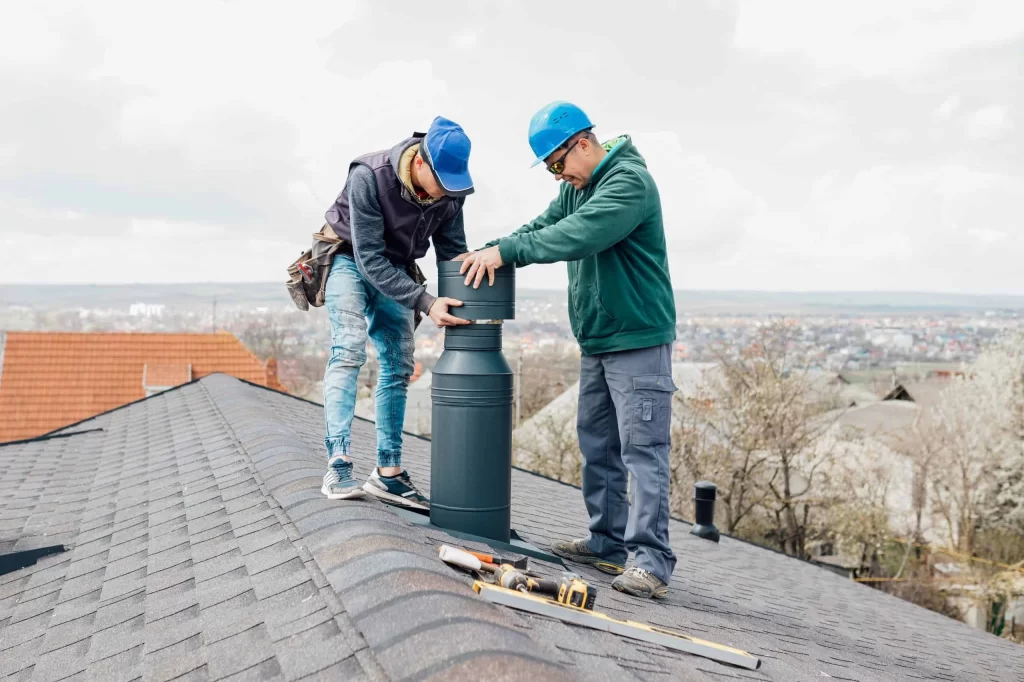 Two workers wearing helmets and safety gear install a metal chimney or vent pipe on a shingled rooftop, tools lying nearby, with a scenic view of houses and trees—showcasing professional air duct services New Jersey residents can trust.