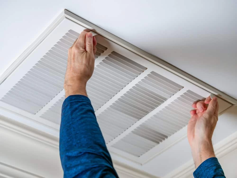 A person wearing a blue long-sleeve shirt is opening or removing a white air vent cover from the ceiling, possibly as part of air duct services New Jersey for improved indoor air quality.