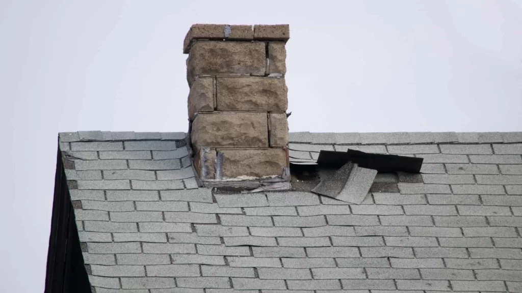 A brick chimney on a gray shingled roof with visible damage; several shingles are lifted and displaced near the chimney, exposing the underlying roofing materials—issues that may also affect air duct services New Jersey homeowners rely on.