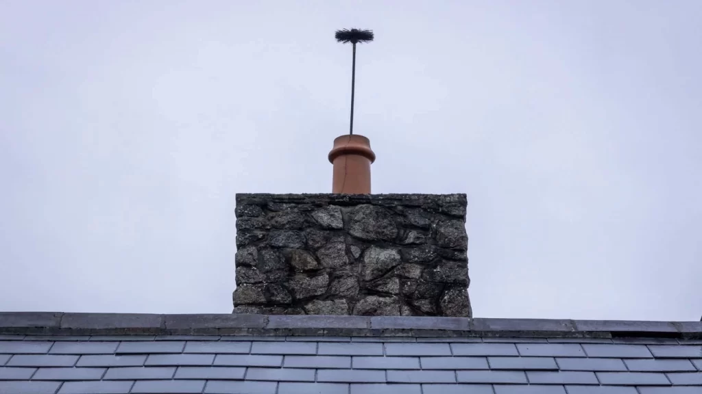 A stone chimney with a terracotta pipe and chimney brush atop, set against a cloudy, gray sky, viewed from behind a slate-tiled roof—a scene evoking the need for expert dryer vent cleaning New Jersey homeowners trust.