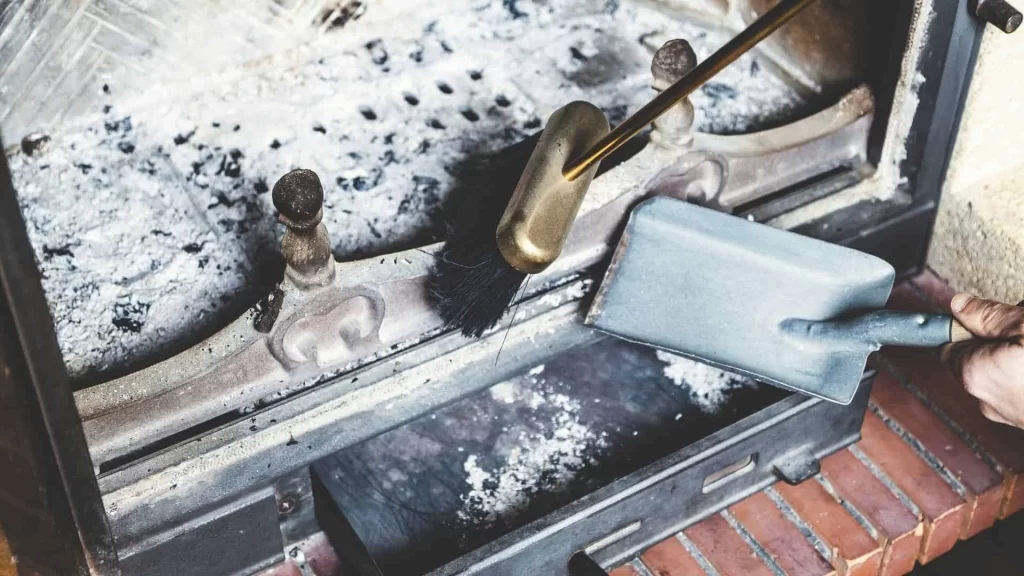 A person cleaning out ashes from a fireplace using a metal dustpan and brush, with gray ash and soot visible inside the fireplace—reminding homeowners of the importance of regular dryer vent cleaning New Jersey for a safer, cleaner home.