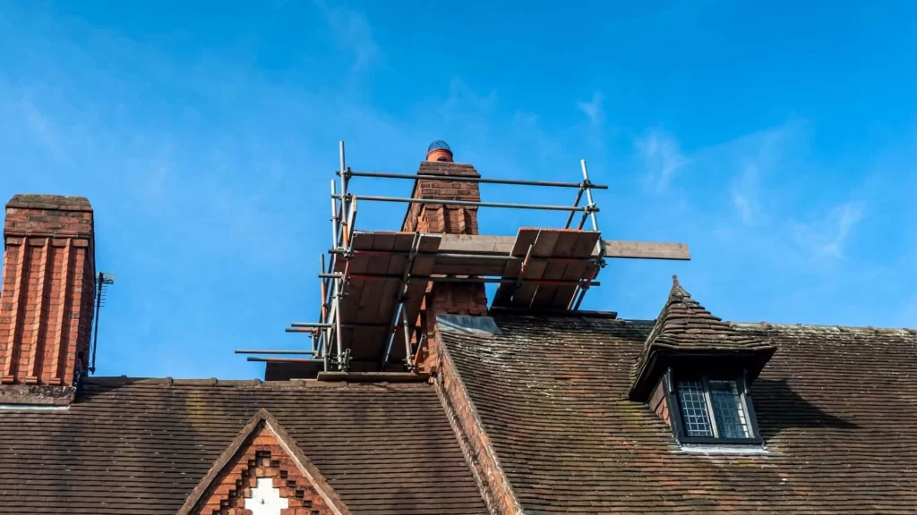 Scaffolding surrounds a brick chimney on a sloped roof under a clear blue sky, indicating construction or repair work—possibly including air duct services New Jersey residents might need for their homes.