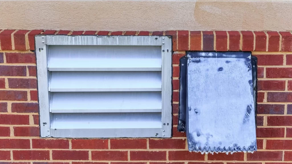 A red brick wall with a beige trim features a silver metal vent—ideal for dryer vent cleaning New Jersey experts—on the left and a frosted or weathered rectangular panel on the right.