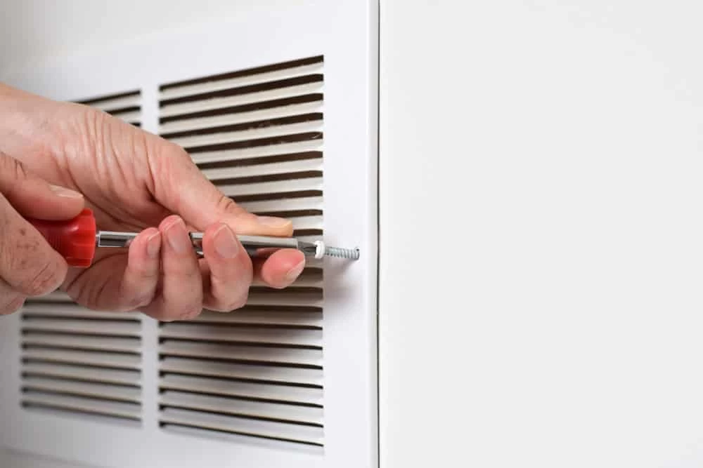 A person uses a screwdriver to remove a screw from a white air vent cover mounted on a wall, an important step in dryer vent cleaning New Jersey.