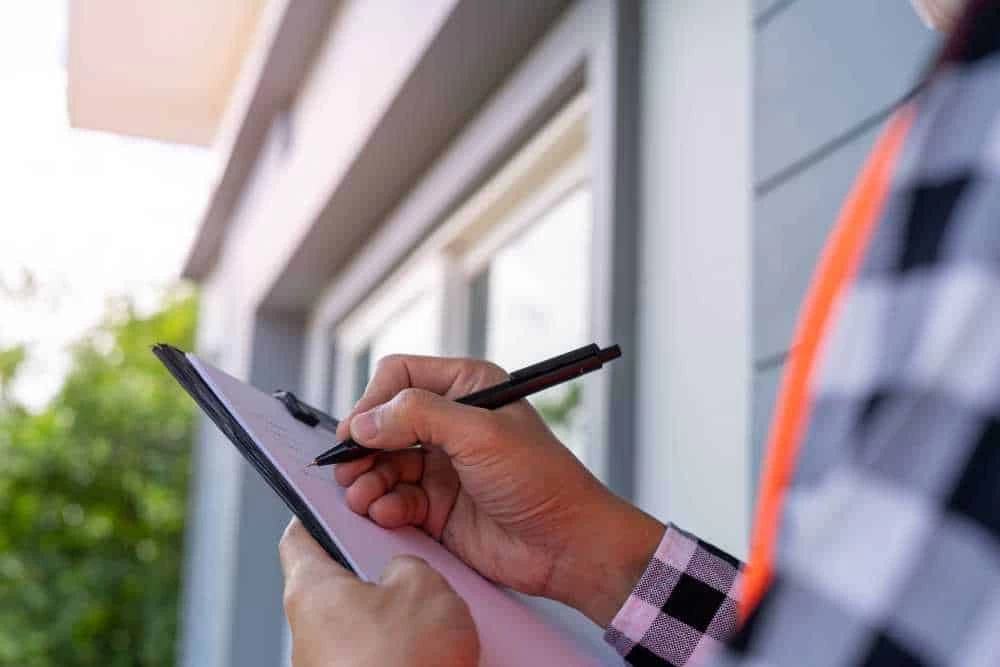A person holding a clipboard and writing with a pen, standing outside near a house with grey siding and windows, wearing a plaid shirt, possibly inspecting for air duct services New Jersey.