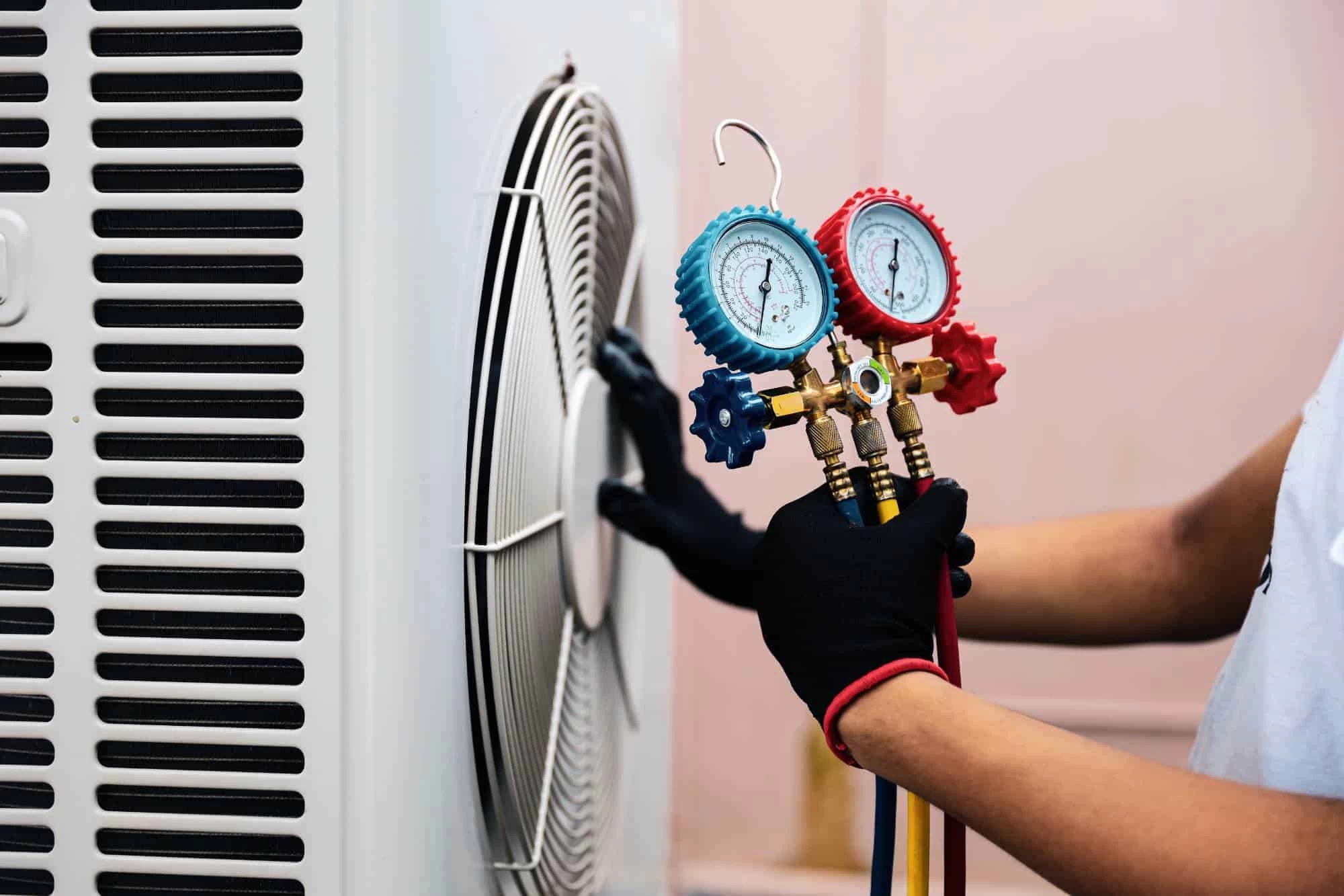 A person wearing black gloves uses a manifold gauge set to check or repair an air conditioning unit, holding the gauges near the unit's fan.