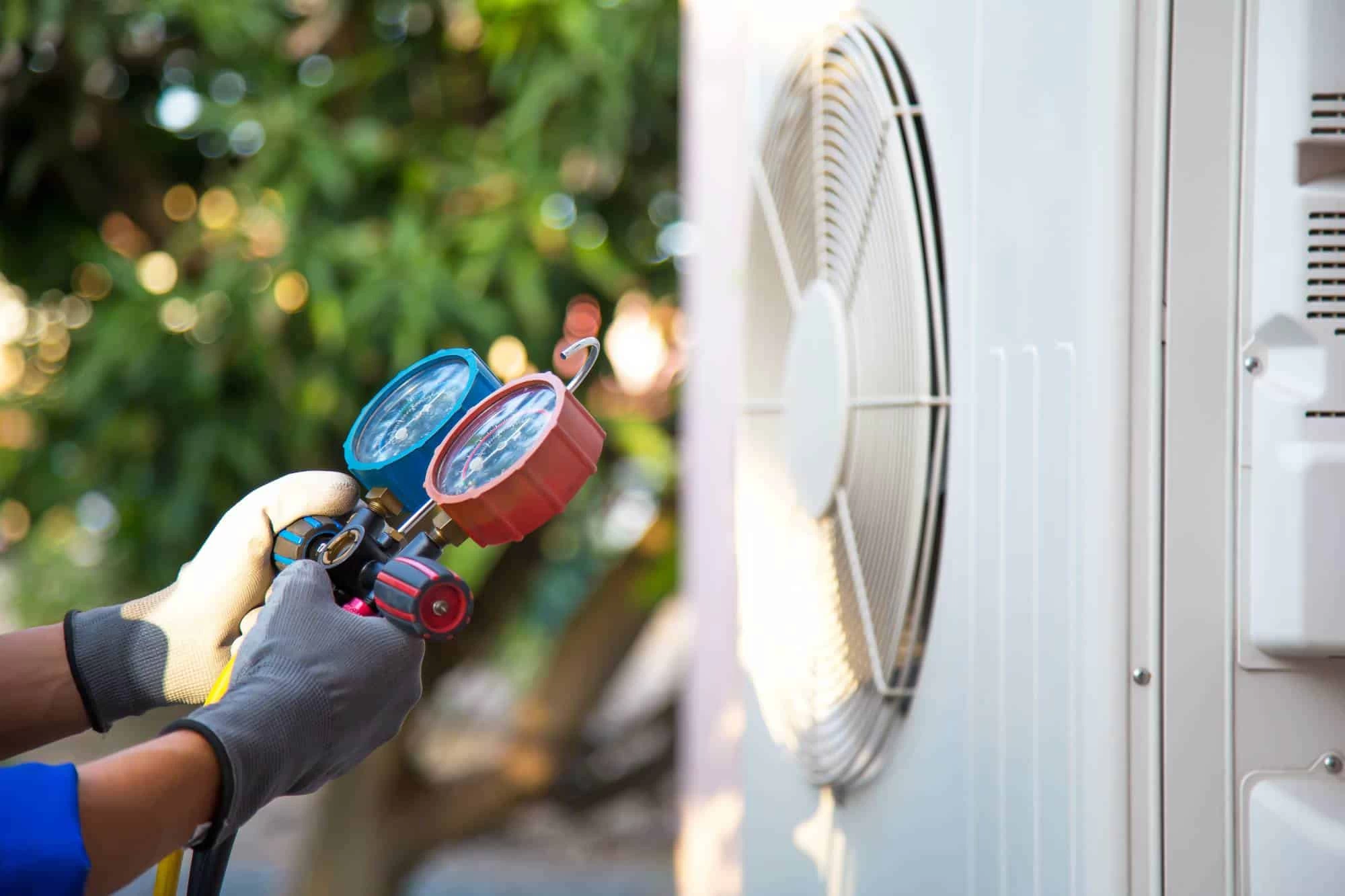 A technician wearing gloves uses a manifold gauge to check the refrigerant pressure of an outdoor air conditioning unit.