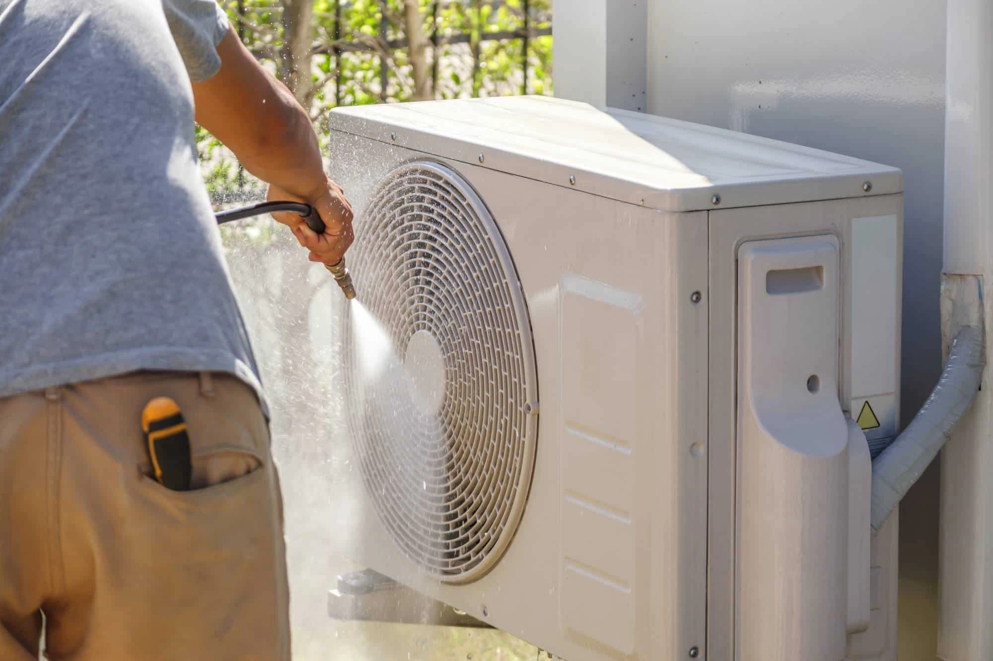 A person wearing a gray shirt and brown pants uses a high-pressure water hose to clean the outdoor unit of an air conditioner. Green foliage is visible in the background.