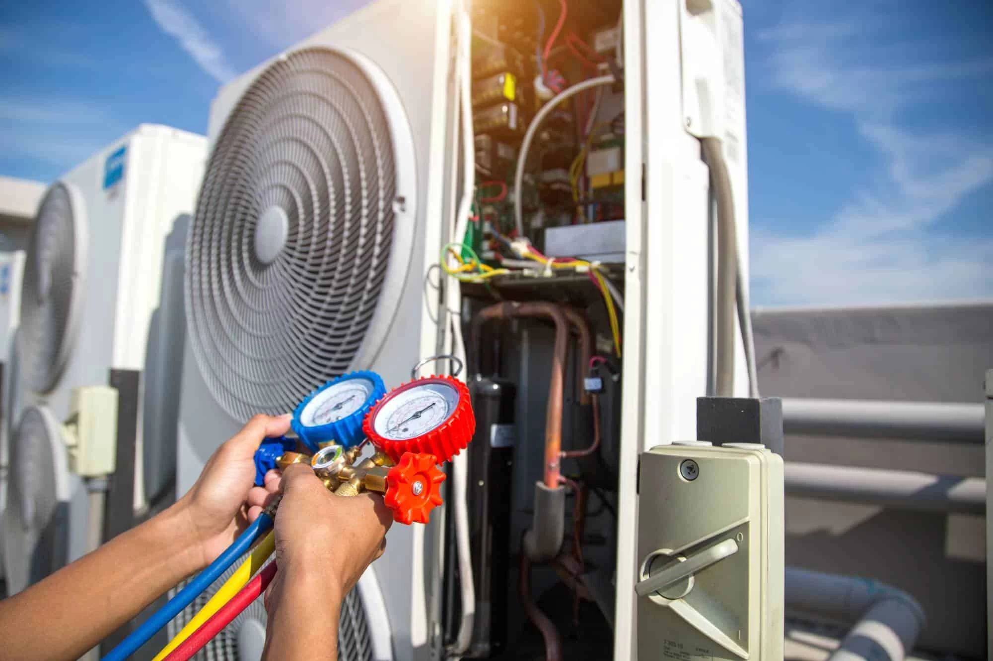 A person uses manifold gauge tools to check the pressure of an outdoor air conditioning unit, with exposed wiring and components visible and another AC unit in the background.