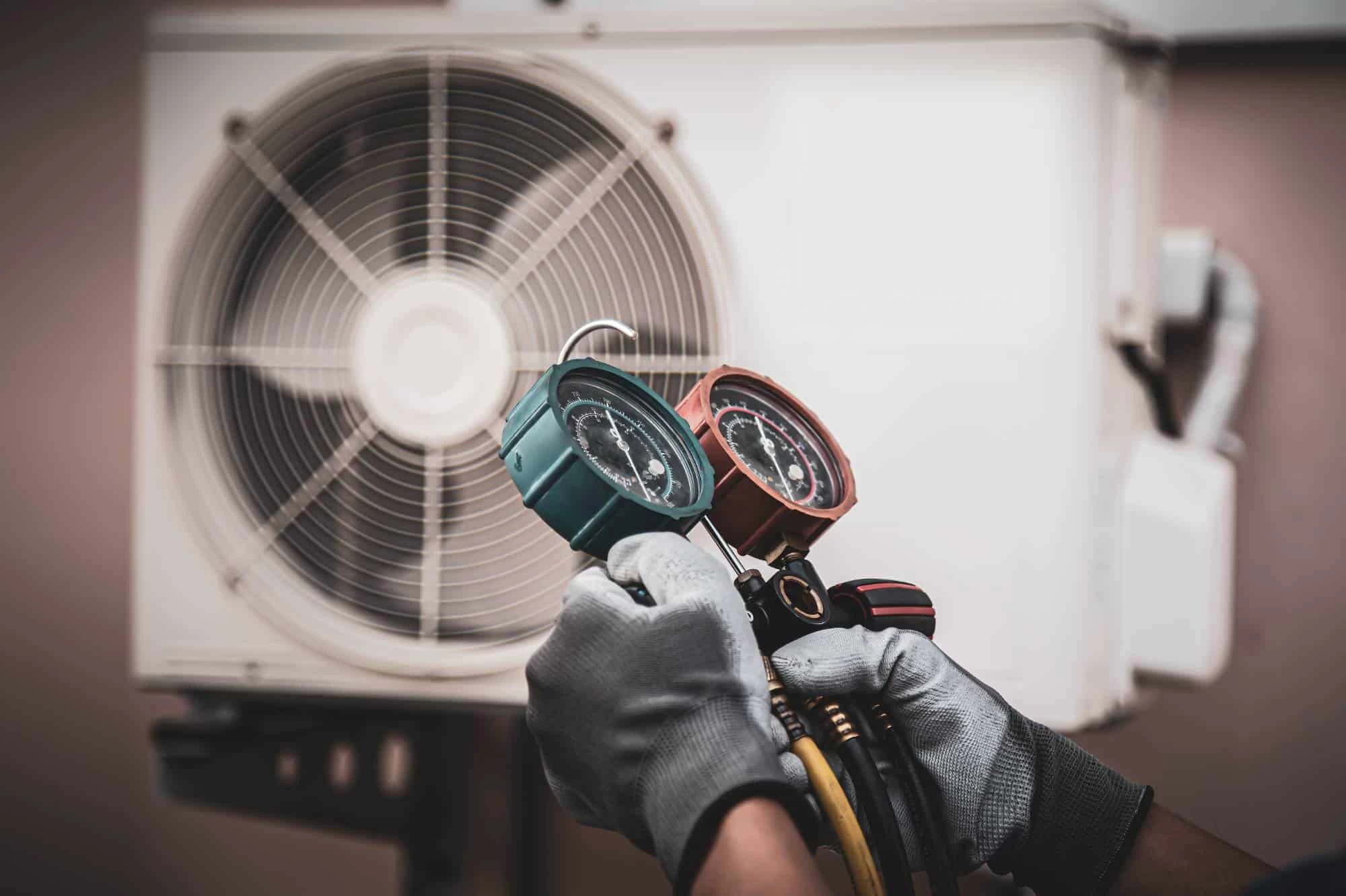 A technician wearing gray gloves uses a manifold gauge set to check a wall-mounted air conditioning unit's refrigerant levels. The fan unit is visible in the background.