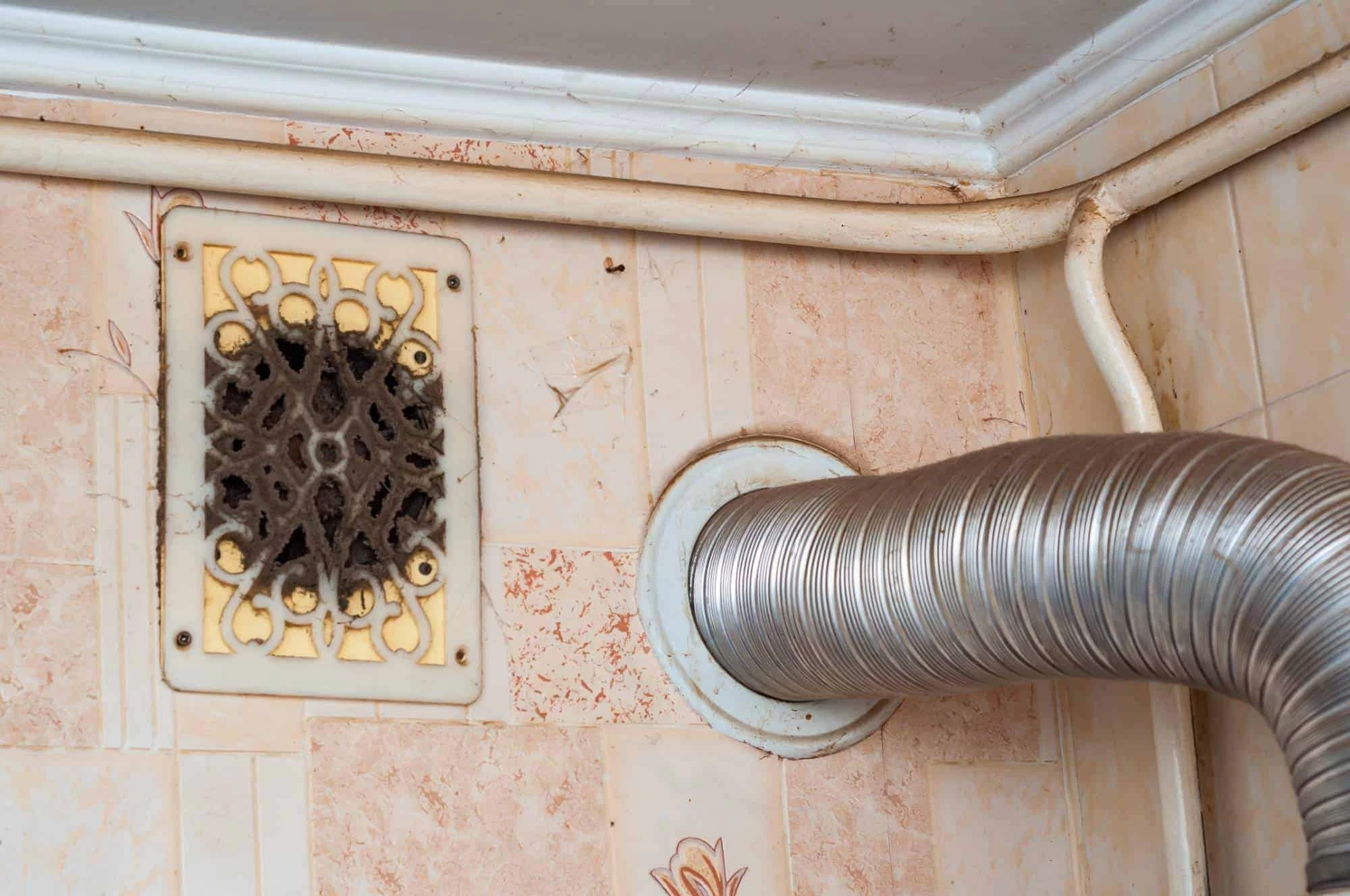 A silver ventilation duct is attached to a wall vent in the corner of a tiled room; an old, ornate, and dusty grill is mounted nearby with visible cobwebs.