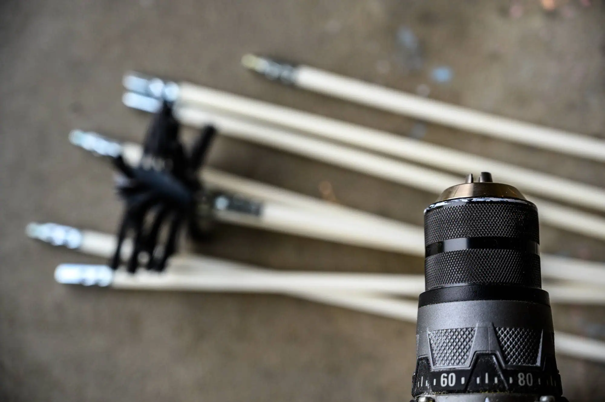 A close-up of a power drill in the foreground with several long rods, possibly cleaning rods, lying on a concrete surface in the blurred background.