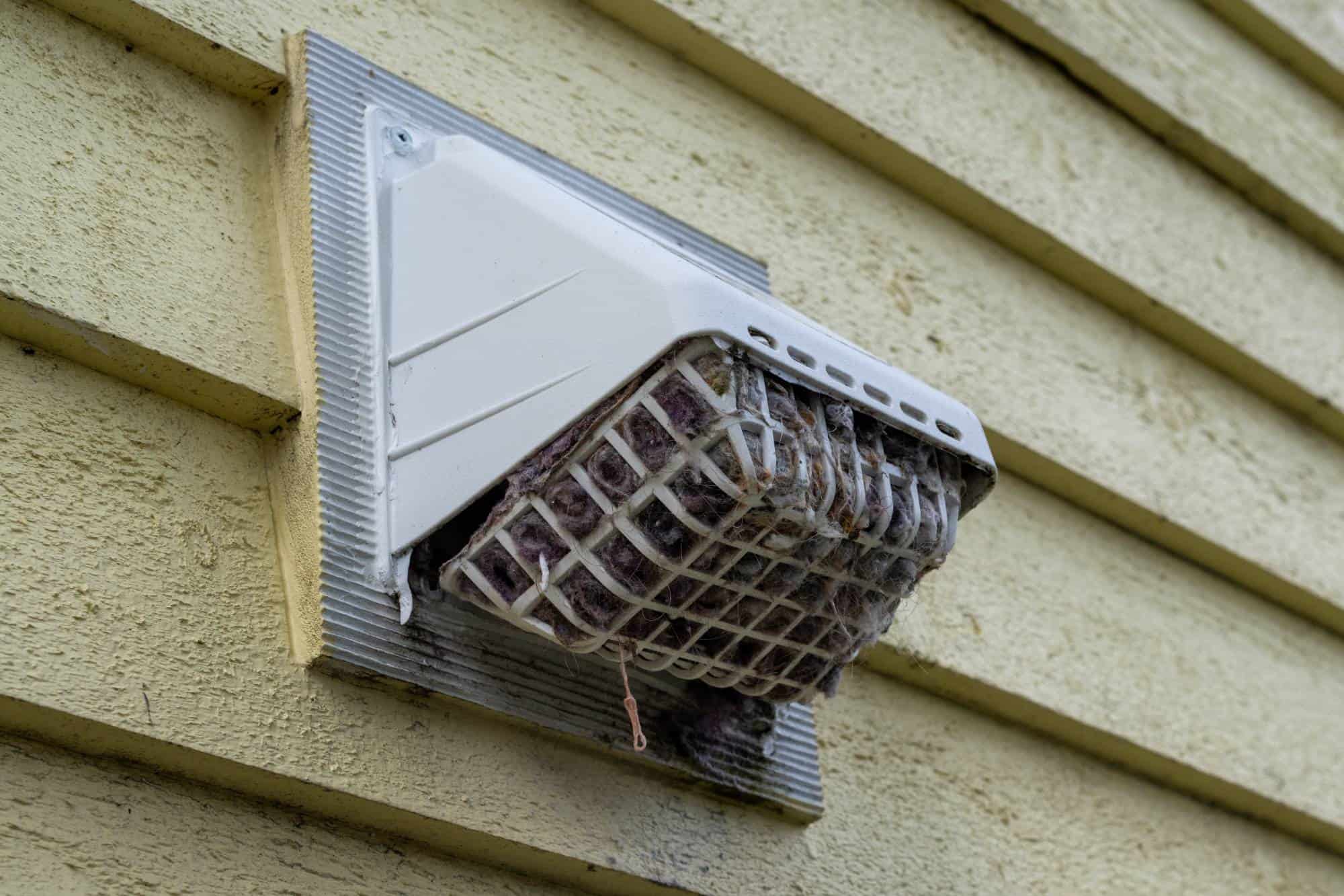 A close-up of a white dryer vent with a plastic cage on the side of a yellow wooden house. The vent appears clogged with lint and debris, posing a potential fire hazard.