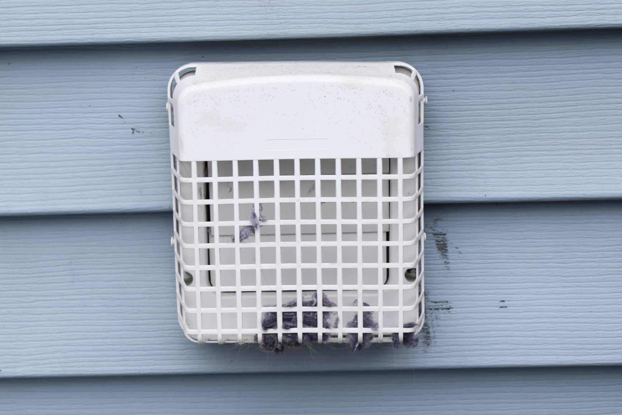 A white vent with a protective grid cover attached to blue siding, partially clogged with lint trapped at the bottom of the grid.