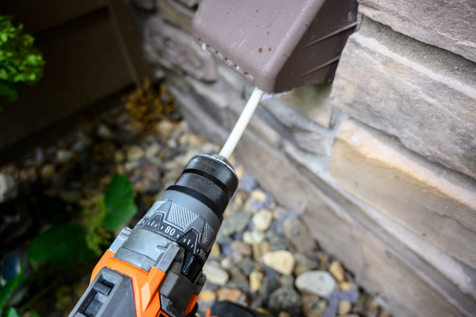 A drill with an orange and black handle is drilling into the wall of a house near an outdoor electrical box, with pebbles and green plants visible on the ground below.