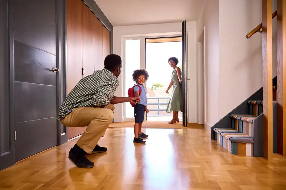 A man kneels on the floor, holding a young child's hand in a bright hallway. A woman stands by the open front door, smiling at them. Stairs are visible on the right, and sunlight streams in—perfect for families seeking dryer vent cleaning New Jersey.