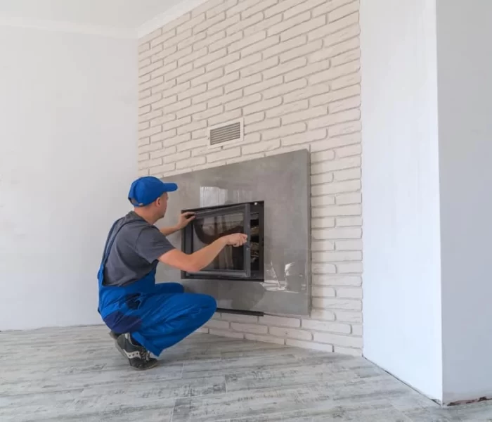 A person in blue overalls and a cap installs or repairs a built-in oven in a white brick wall inside a modern, unfinished room—services like air duct cleaning New Jersey can also help keep home appliances running efficiently.
